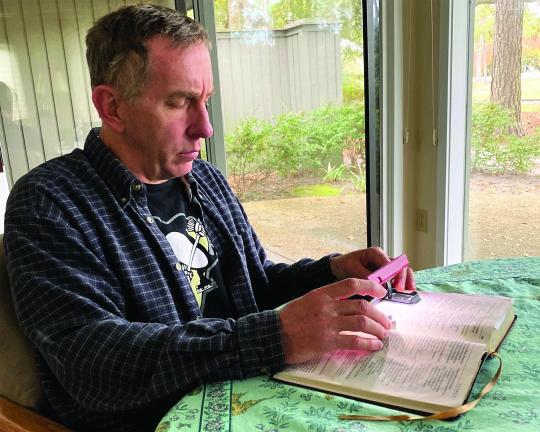 Don sitting at the table reading a book with a magnifier