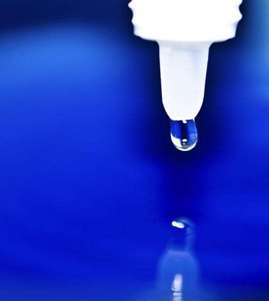 eyedrops bottle suspended over blue watery background with one drop showing; reflection of bottle appears underneath