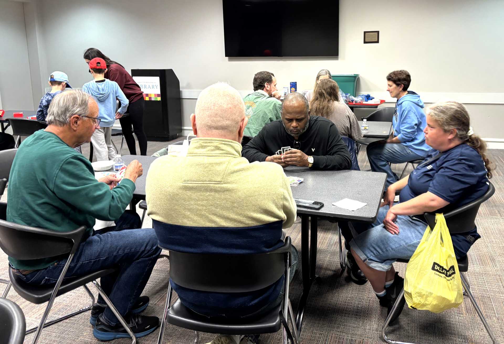 Small groups of people play board games and cards at tables in a library community room.