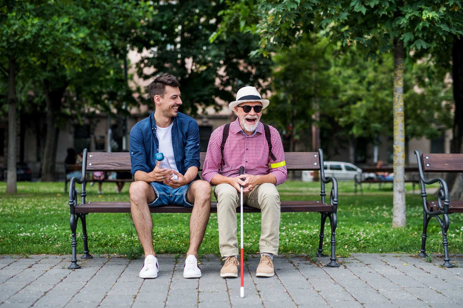 Two men sitting and talking on a bench, younger man on left wearing blue shirt older man sitting on right wearing a fedora and holding a white cane